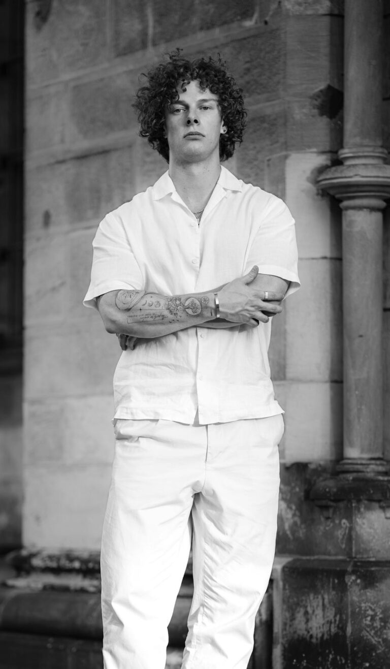 Black and white portrait of a stylish man in white attire, standing confidently outdoors.